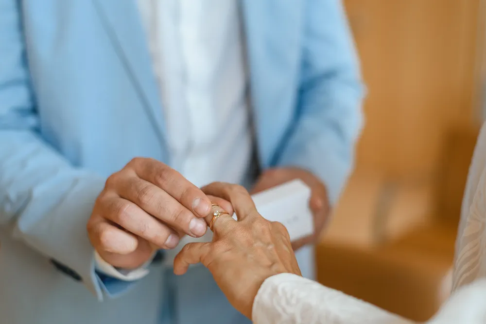 Man in light blue suit presents ring to woman in white garment during intimate indoor ceremony.