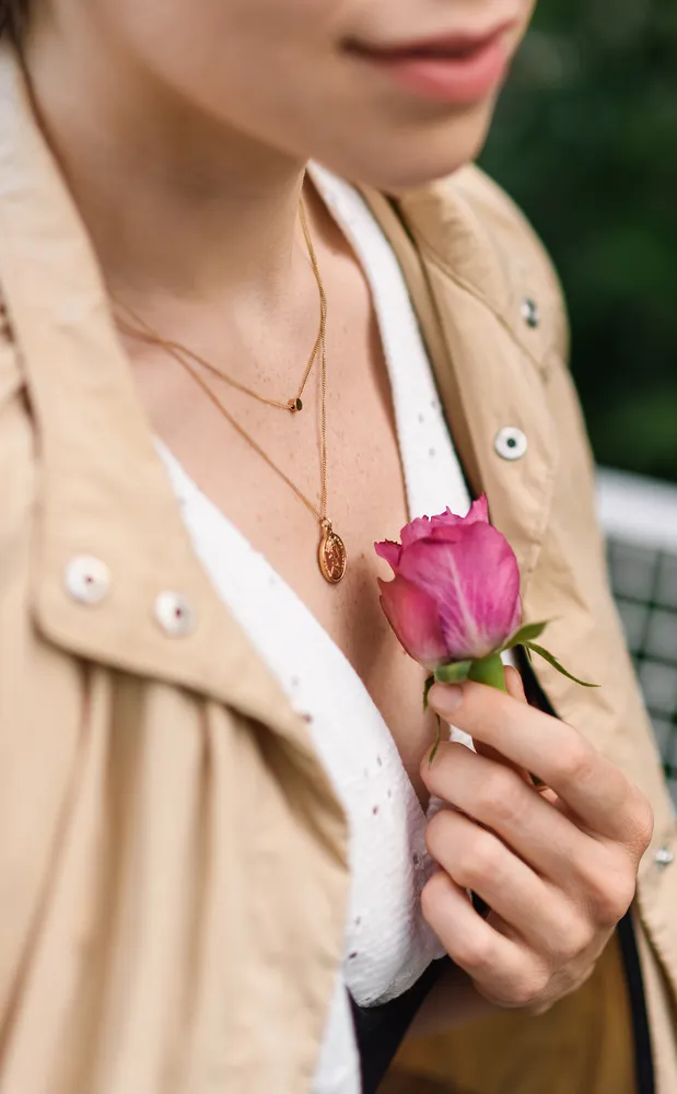 A woman in a tan jacket holds a pink rose against a green backdrop, her gold necklace with a pendant visible.