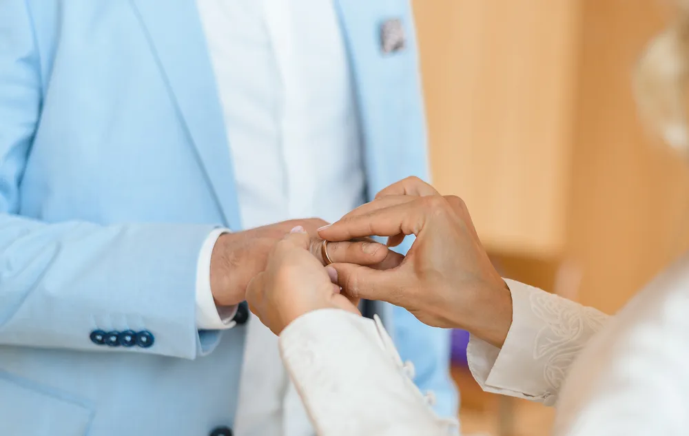 A person in a light blue suit receives a gold ring from another individual in a white embroidered garment, suggesting a marriage proposal or ceremony. The background is softly blurred, focusing on the hands and ring exchange.