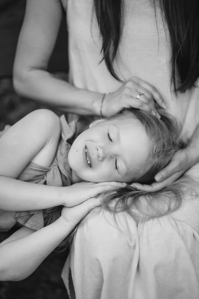 A woman in a light-colored dress gently cradles a sleeping child's head on her lap. The child wears a ruffled dress, eyes closed in peaceful slumber. Both are in monochrome, creating a timeless, serene portrait of care and restfulness.
