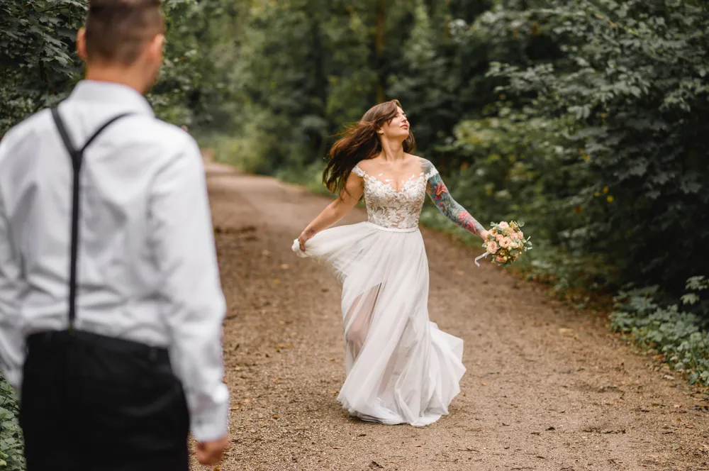 A woman in a white lace wedding dress twirls joyfully on a dirt path surrounded by dense greenery, holding a bouquet, with a man in a white shirt and black pants walking behind her.