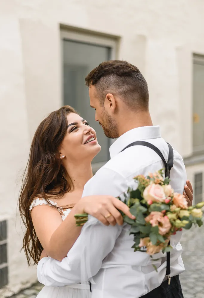 Couple embracing; woman in white dress holds peach roses, man in white shirt and suspenders, cobblestone street, white building background.