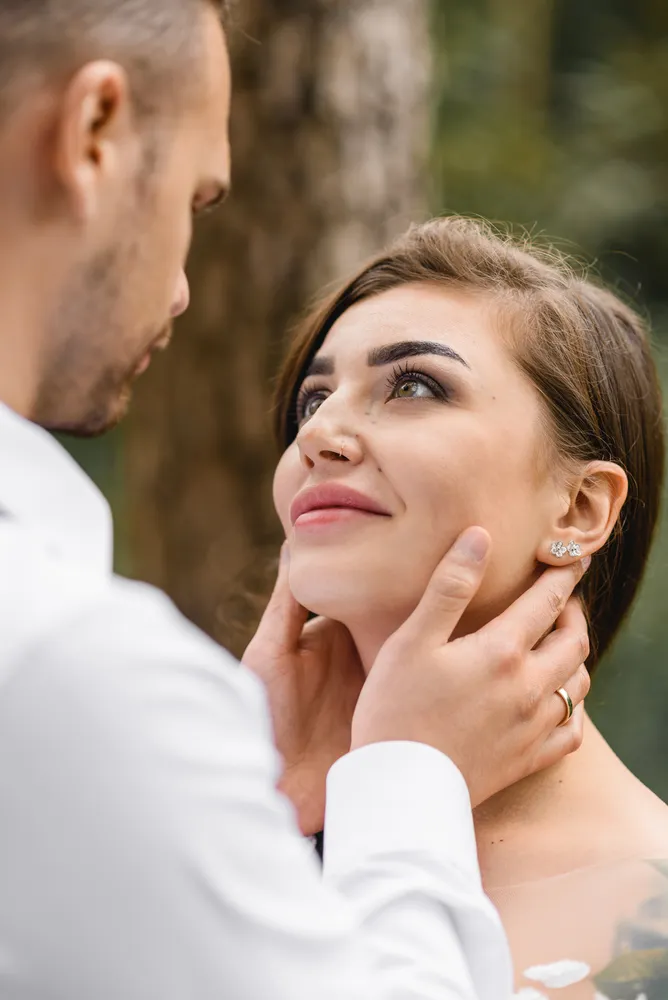 Man in white shirt gently touches woman's face. She smiles, wearing diamond earrings. Outdoors, blurred foliage background. Intimate moment captured.