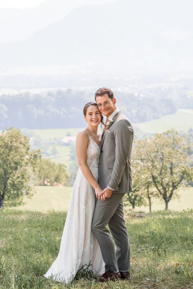 Couple in wedding attire stands in a grassy field, embracing, with a scenic landscape in the background.