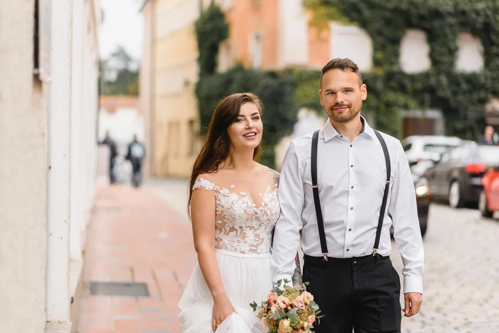 Couple in wedding attire strolls down cobblestone street, woman holds bouquet, historic buildings in background.