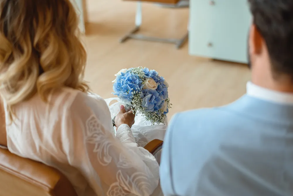 A woman with blonde hair holds a blue and white bouquet, seated beside a man in a light blue suit, in a softly lit room with wooden floors.