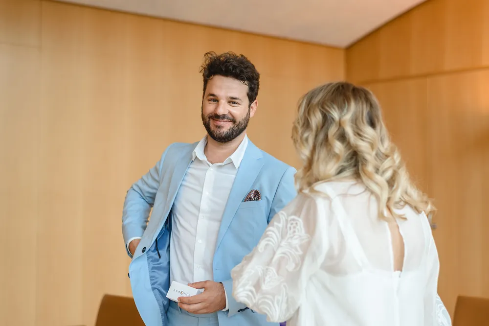 Man in light blue suit smiles, holding card, conversing with woman in white dress. Both in room with wooden walls. Professional attire suggests formal event.
