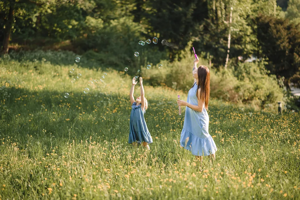 A woman and a young girl in light blue dresses play with soap bubbles in a sunlit meadow surrounded by trees and wildflowers. The woman blows bubbles while the girl reaches up to touch them.