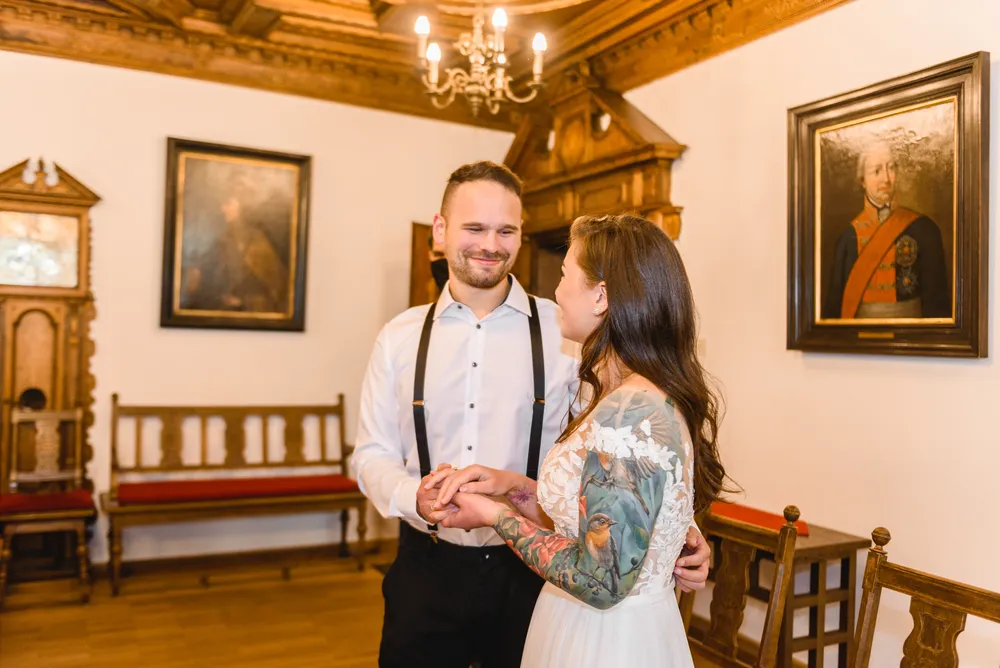 A smiling couple, hands clasped, in a historic room with wooden furniture and classical portraits, wearing formal attire, suggestive of a wedding or formal event.