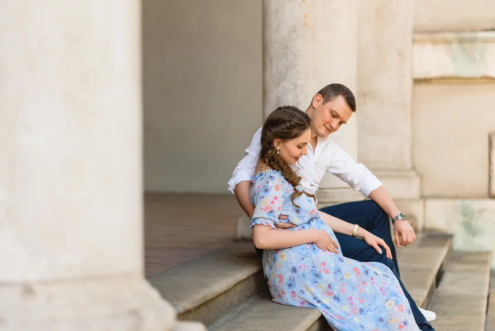 Couple seated on steps of a historic building. Woman in floral dress cradles her belly, man in white shirt smiles. Warm, natural lighting. Architectural columns and stone steps frame the scene.