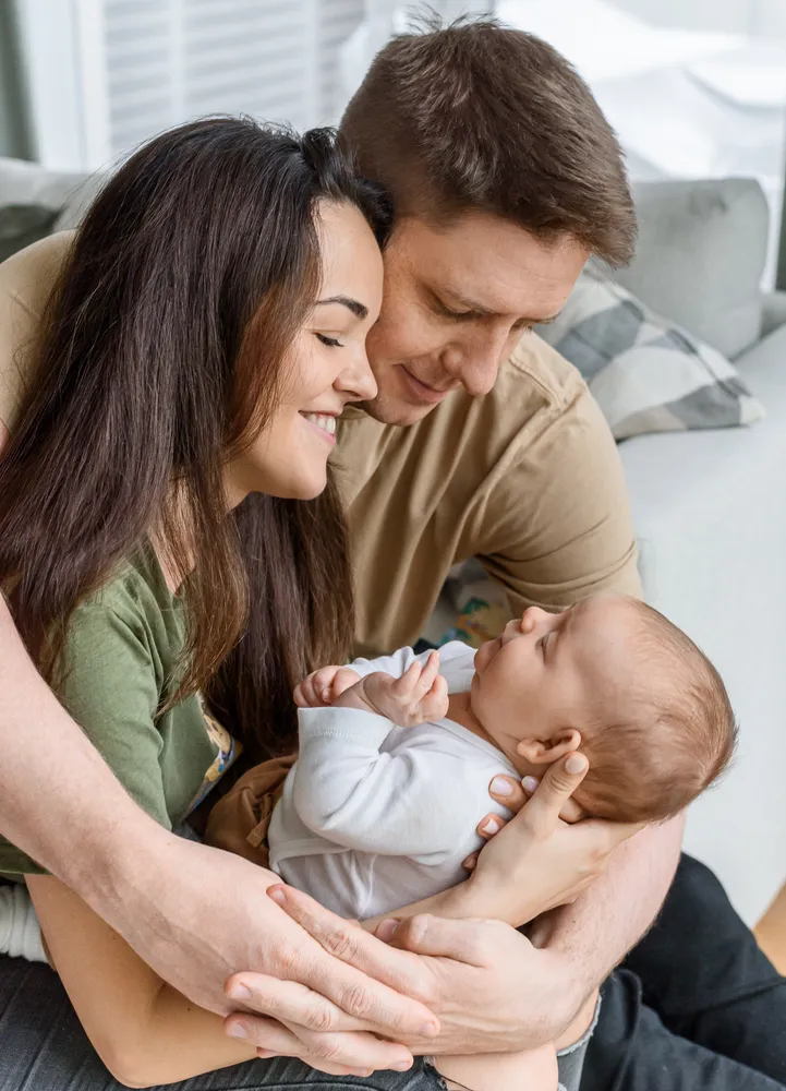 Family of three shares a tender moment indoors, mother and father cradling sleeping infant on a couch, all smiling.