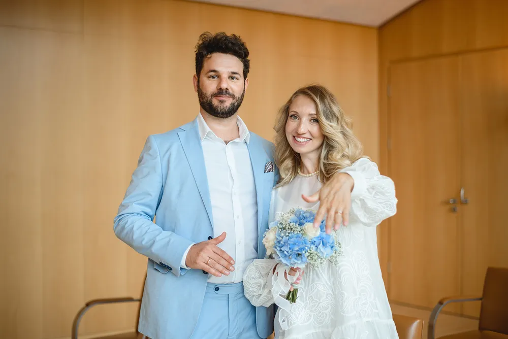 A man and woman in wedding attire stand together in an interior space. The man wears a light blue suit, and the woman is in a white lace wedding dress holding a bouquet of blue and white flowers. Both are smiling at the camera.